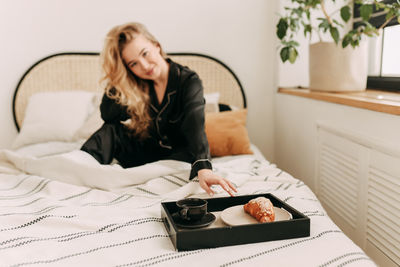A happy pretty young woman in pajamas is resting and having breakfast in bed in a cozy apartment