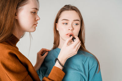 Close-up of woman applying lipstick on beautiful young friend over white background