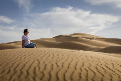 Woman sitting on sand dune at desert against sky