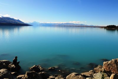 Rear view of man standing on rock by calm lake against clear blue sky