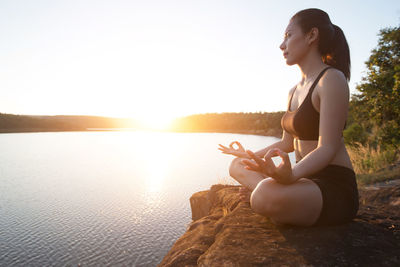 Young woman practicing yoga in lotus position while sitting on rock at lakeshore