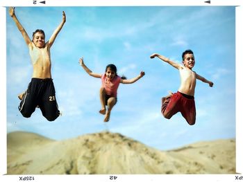 Portrait of cheerful siblings jumping on sand against blue sky