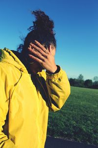 Side view of woman wearing yellow hooded shirt covering face while standing on grassy field against clear blue sky