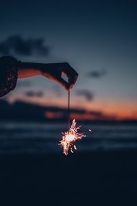 Cropped hand holding burning sparkler against sky during sunset