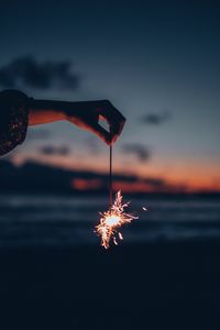 Hand holding illuminated fireworks against sky during sunset