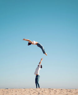 Full body side view of sporty female acrobat performing somersault high in air against blue sky over man standing on sandy seashore