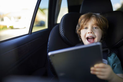 Portrait of happy boy using tablet pc while sitting at back seat in car