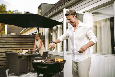 Smiling mature man preparing barbecue meal while in woman in background at back yard