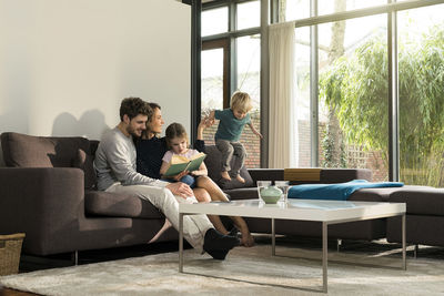 Family on sofa at home reading book with boy jumping