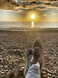 Low section of person on pebbles at beach against sky during sunset