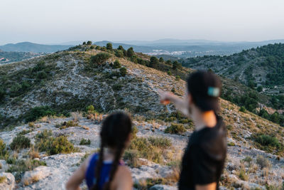 Back view of unrecognizable young man pointing away while standing with female friend on hill and observing environment in highlands during hiking trip together