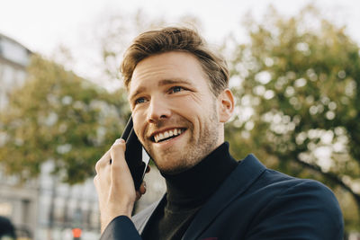 Smiling businessman talking through smart phone while looking away in city