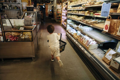 Rear view of boy holding basket while running near refrigerated section in supermarket