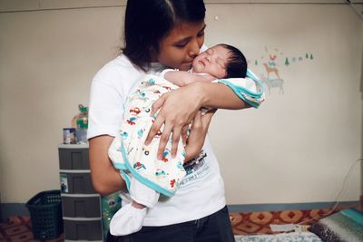 Mother kissing cute daughter while standing against wall at home