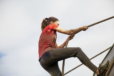 Low angle view of woman climbing wall with rope against cloudy sky