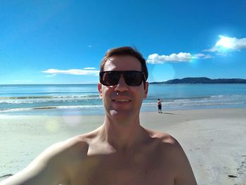 Smiling shirtless man wearing sunglasses at beach against sky on sunny day