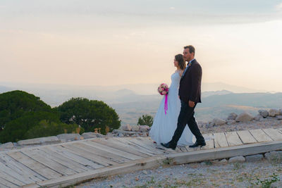 Side view of bride and groom walking on boardwalk against sky