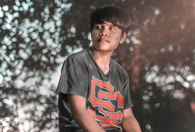 Young man looking away while standing against trees
