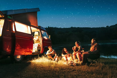 Cheerful group of friends having fun camping near lake and van during starry night