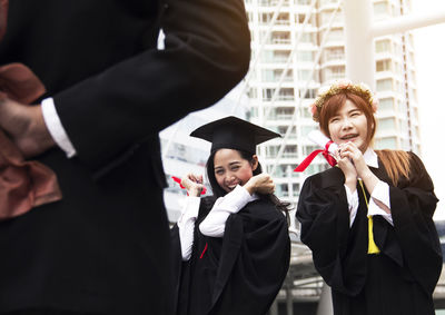 Happy friends in graduation gown standing in city