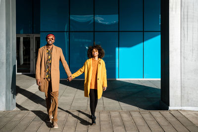 Full body of cheerful african american man and woman in stylish beige and yellow coats holding hands on street looking at camera