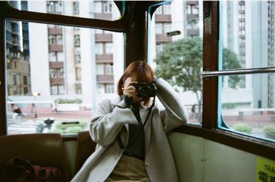 Woman photographing with camera while sitting by window in tram