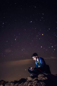 Young man using phone while sitting on rock against star field