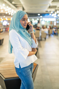 Young woman wearing hijab talking on smart phone while standing in restaurant