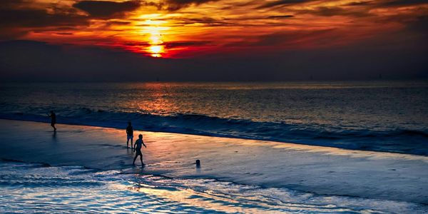 Silhouette people walking on beach against orange sky