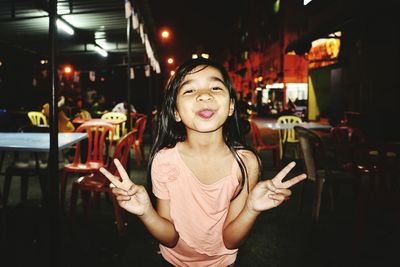 Portrait of girl sticking out tongue while gesturing peace sign in city at night