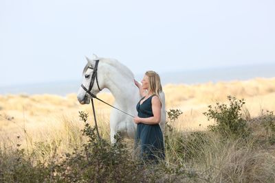 Side view of woman stroking white horse while standing on land against sky