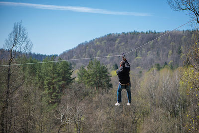 Rear view of man hanging on zip line by trees at forest