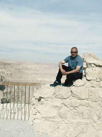 Man wearing sunglasses while sitting on rock against sky
