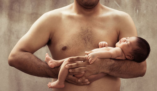 Midsection of shirtless father carrying son against wall