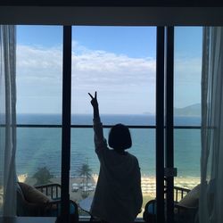 Rear view of woman gesturing peace sign while looking at sea through window at home