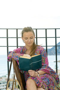 Pretty young woman sitting on a terrace by the sea reading a book