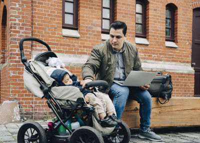 Father using laptop while son sitting in stroller against building in city