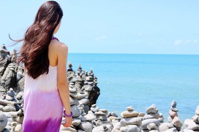 Rear view of young woman standing on stones by sea against sky