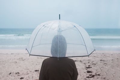 Back view of unrecognizable traveler in raincoat with transparent umbrella walking on empty sandy beach towards waving sea in cloudy day