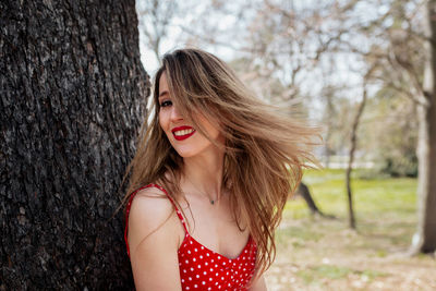 Portrait of smiling woman standing against tree trunk in park