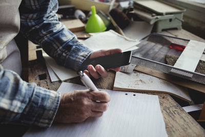Cropped image of owner holding mobile phone while writing on paper at workbench