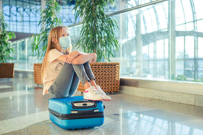 Full length of girl wearing mask sitting on suitcase at airport