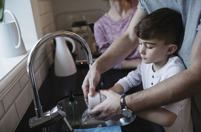 Midsection of man assisting boy in washing dishes by woman in kitchen