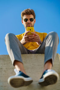 Low angle view of young man using phone sitting against sky