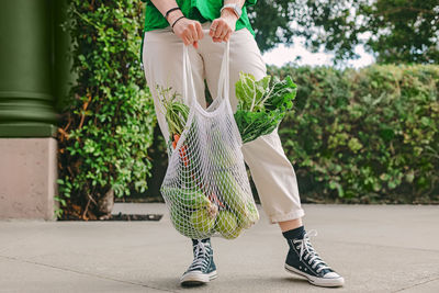 Unrecognizable woman in green shirt holding a mesh bag with veggies, standing on the street