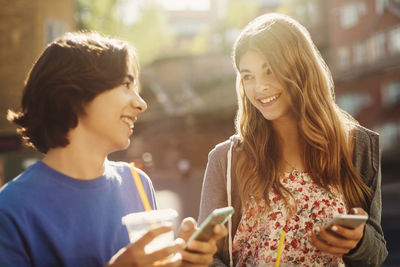 Happy teenagers looking at each other while holding smart phones outdoors