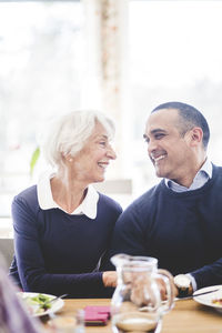 Cheerful senior man sitting face to face with son while having lunch at table in nursing home