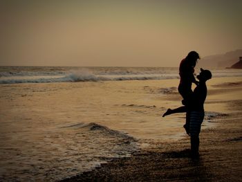 Side view of man lifting woman while standing at beach against sky during sunset