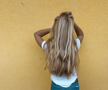 Rear view of young woman standing with hand in hair against yellow wall
