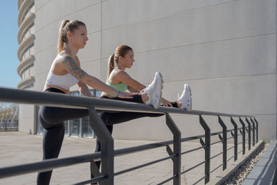 Side view of determined young slim women in sportswear doing stretching exercise for legs while standing near metal railing during outdoor fitness workout in city